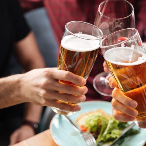 Cropped photo of young friends sitting in cafe while drinking alcohol. Focus on glasses of beer.
